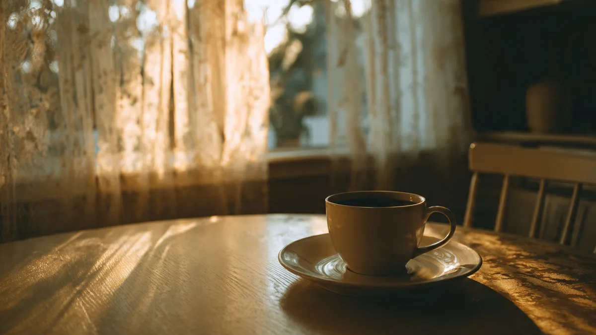 A quiet kitchen table at dawn with a single empty plate and a cup of black coffee, warm morning light streaming through a window