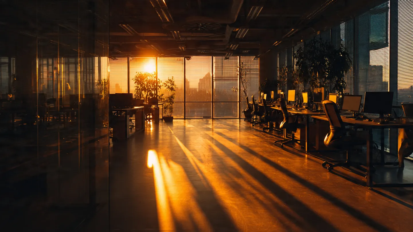 Empty office desks with morning light streaming through tall windows, chairs pushed back as if recently abandoned